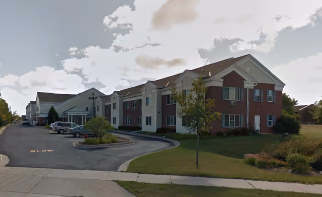Exterior view of a two-story senior living facility building with a brick and white siding facade, a driveway with parked cars, a small landscaped area with trees and shrubs, and a partly cloudy sky.