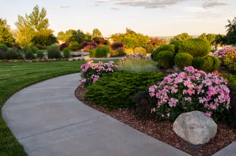 A curved concrete pathway winds through a landscaped garden with green grass, various shrubs, and blooming pink flowers under a partly cloudy sky.