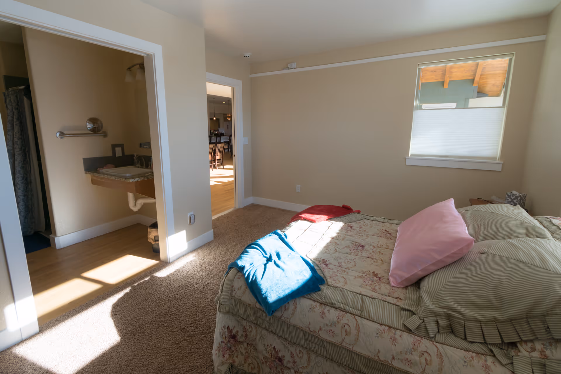 Sunlit bedroom with a floral bed and pillows, a window, and an open doorway leading to a bathroom and hallway.