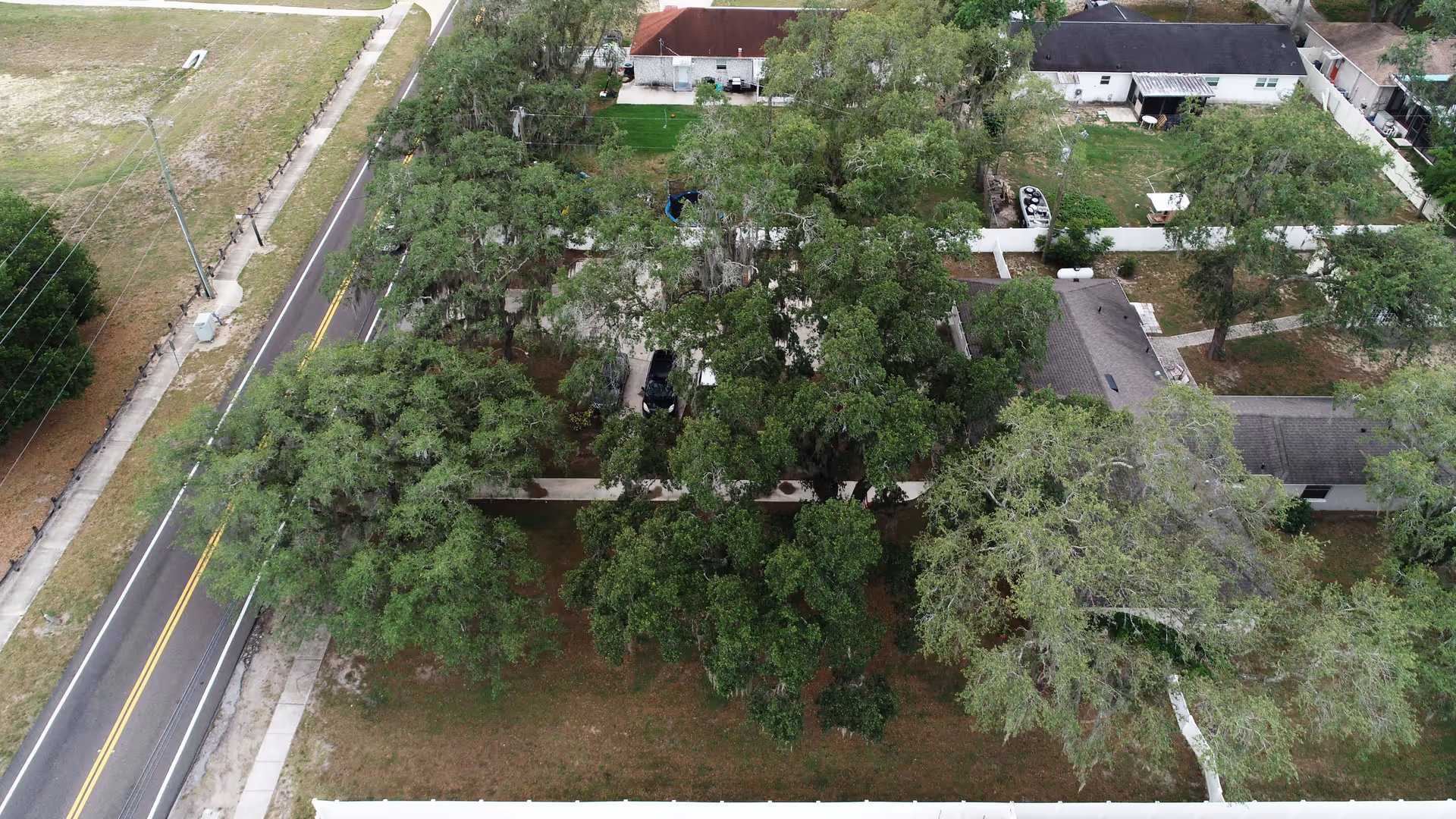 Aerial view of a residential area with several houses surrounded by large green trees. A road runs along the left side of the image with sidewalks on both sides. The houses have yards with grass and some outdoor items like a trampoline and a small boat. The area is fenced in parts and appears quiet and suburban.