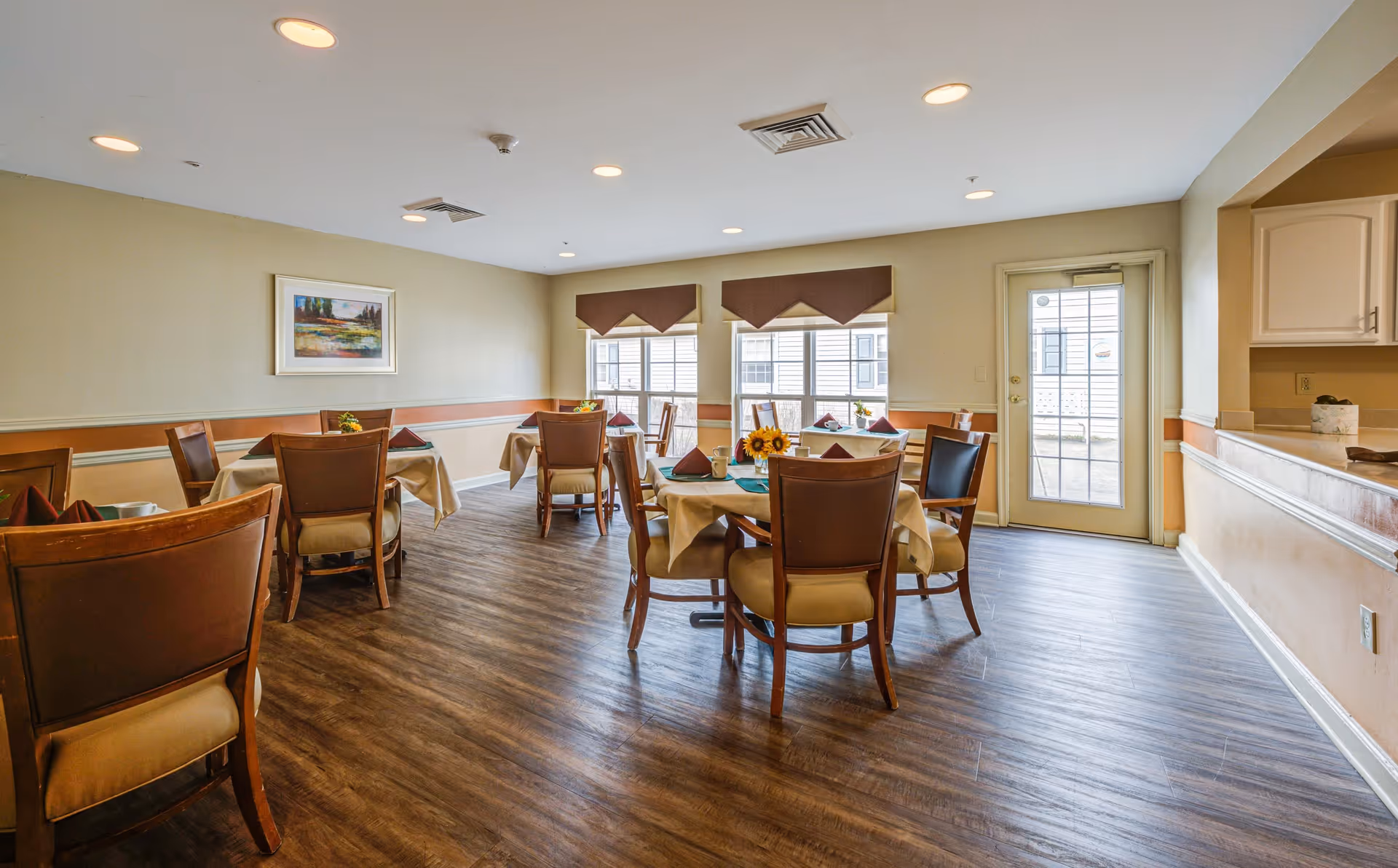 A dining room in a senior living facility with several round tables covered with beige tablecloths, each set with folded napkins and small flower arrangements. The room has wood flooring, beige walls with a chair rail, and windows with brown valances letting in natural light. There is a door with glass panes on the right side and a counter area adjacent to it.