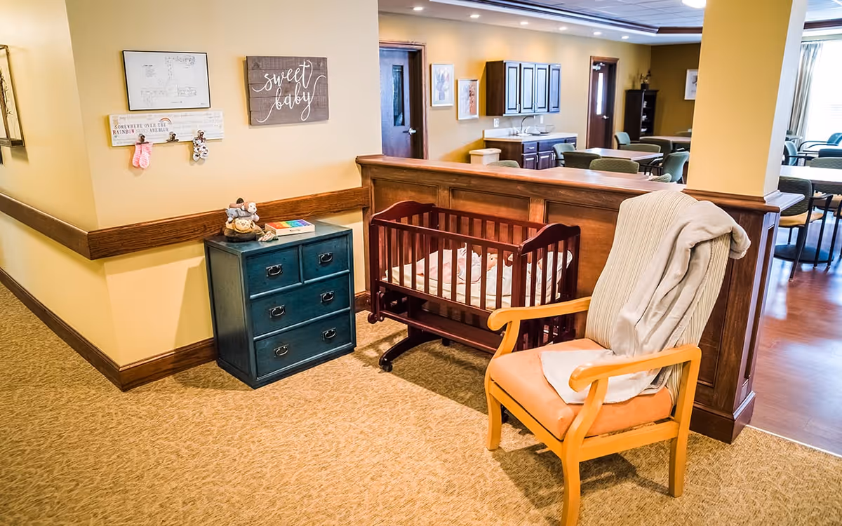 A cozy corner in a senior living facility featuring a wooden baby crib, a cushioned armchair with a blanket draped over it, and a small blue chest of drawers with decorative items on top. The wall behind has framed artwork and a sign that reads 'sweet baby'. In the background, there is a dining area with tables and chairs.
