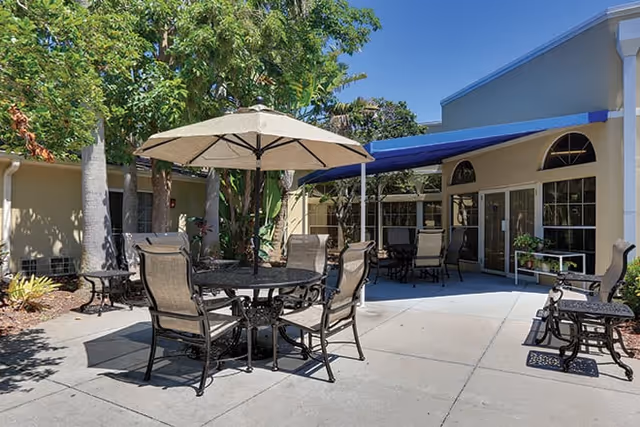 Sunlit outdoor courtyard with round metal tables, umbrella-shaded seating, and patio chairs in front of a single-story building.
