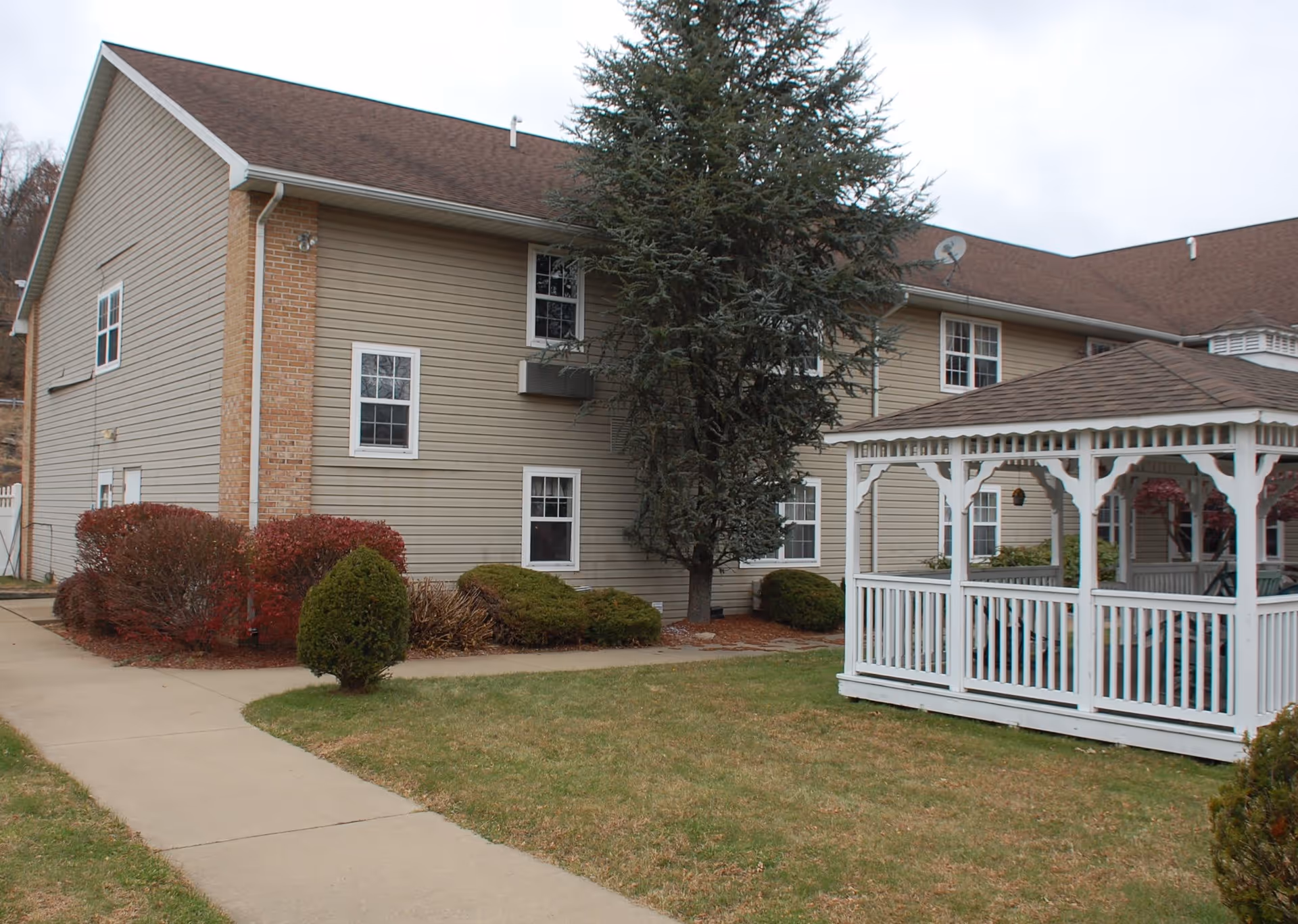 Exterior view of a senior living facility building with beige siding and a brown roof. There is a white wooden gazebo on the right side, surrounded by green grass, bushes, and a tree. A concrete walkway curves through the lawn toward the building.