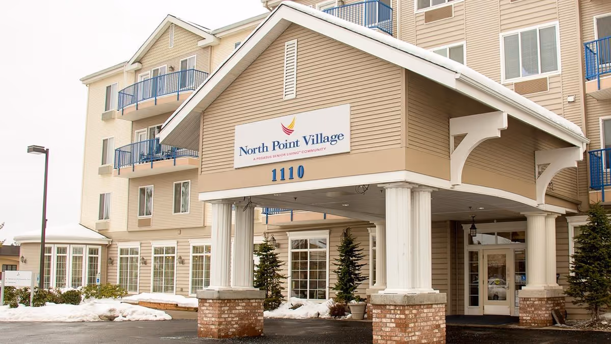 Exterior view of North Point Village senior living community building with a covered entrance supported by white columns. The building has beige siding, multiple windows, and blue balcony railings. Snow is visible on the ground around the entrance.