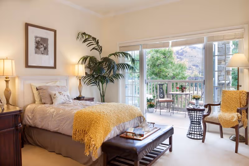 A cozy bedroom with a single bed covered in beige bedding and a yellow throw blanket. There are two bedside tables with lamps, a large potted plant, and a framed picture on the wall. The room has a sliding glass door leading to a balcony with outdoor seating and a view of trees and hills.