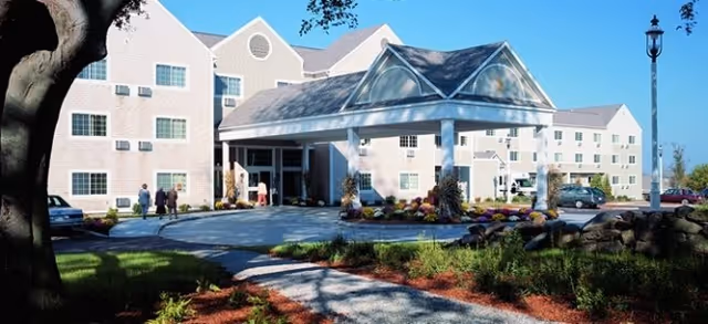 Front exterior of a multi-story senior living building with a covered entrance, driveway, landscaped grounds, and a few people near the entrance.