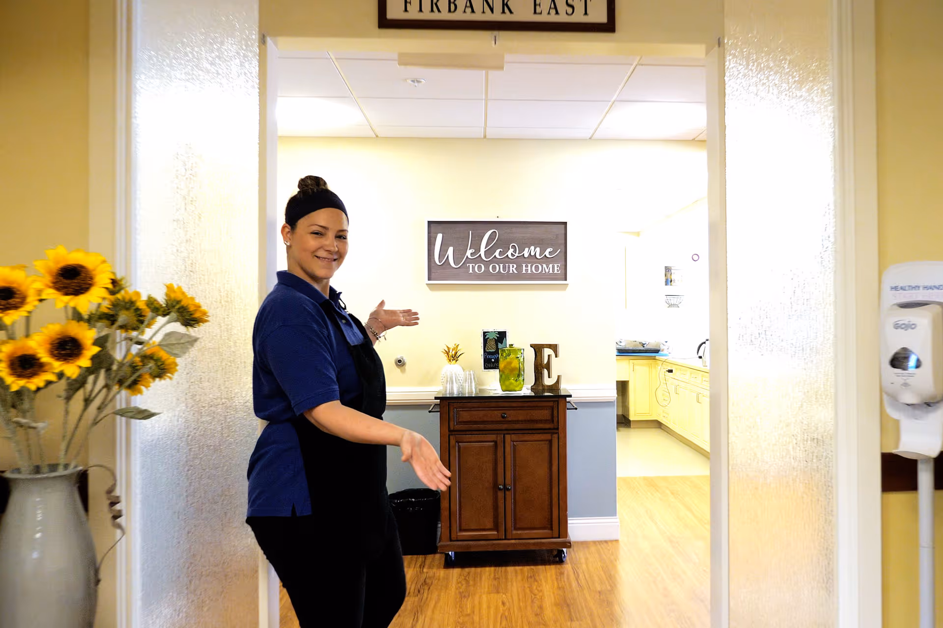 A woman wearing a navy blue shirt and black apron stands in a hallway with wooden flooring, smiling and gesturing towards a room. The room has a wooden cabinet with decorative items on top and a sign on the wall that reads 'Welcome to our home'. There is a vase with sunflowers on the left side of the hallway and a hand sanitizer dispenser mounted on the right wall.