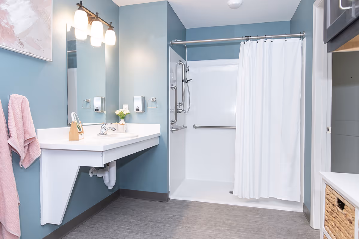 Accessible bathroom with a wall-mounted sink and mirror, lit vanity lights, teal walls, and a grab-bar shower with a white curtain.
