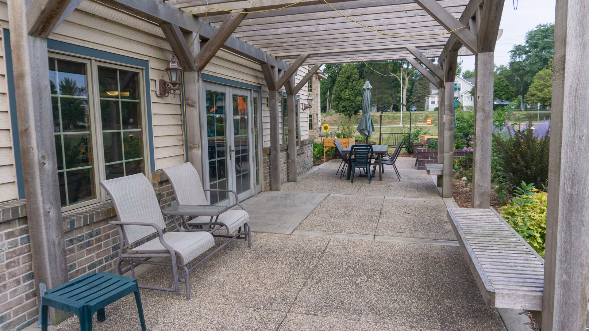 Covered outdoor patio with lounge chairs, a dining table under a wooden pergola next to a building and garden.