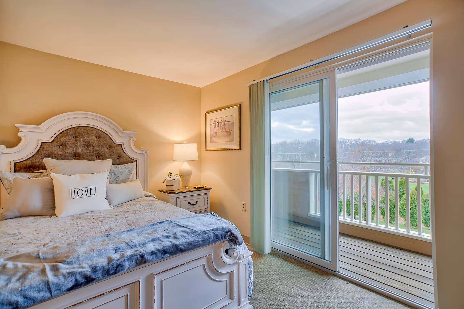A cozy bedroom with a large white wooden bed featuring a tufted headboard and multiple pillows, including one with the word 'LOVE'. There is a nightstand with a lamp and a small decorative item next to the bed. A framed picture hangs on the beige wall. Sliding glass doors open to a balcony with a view of trees and a cloudy sky.