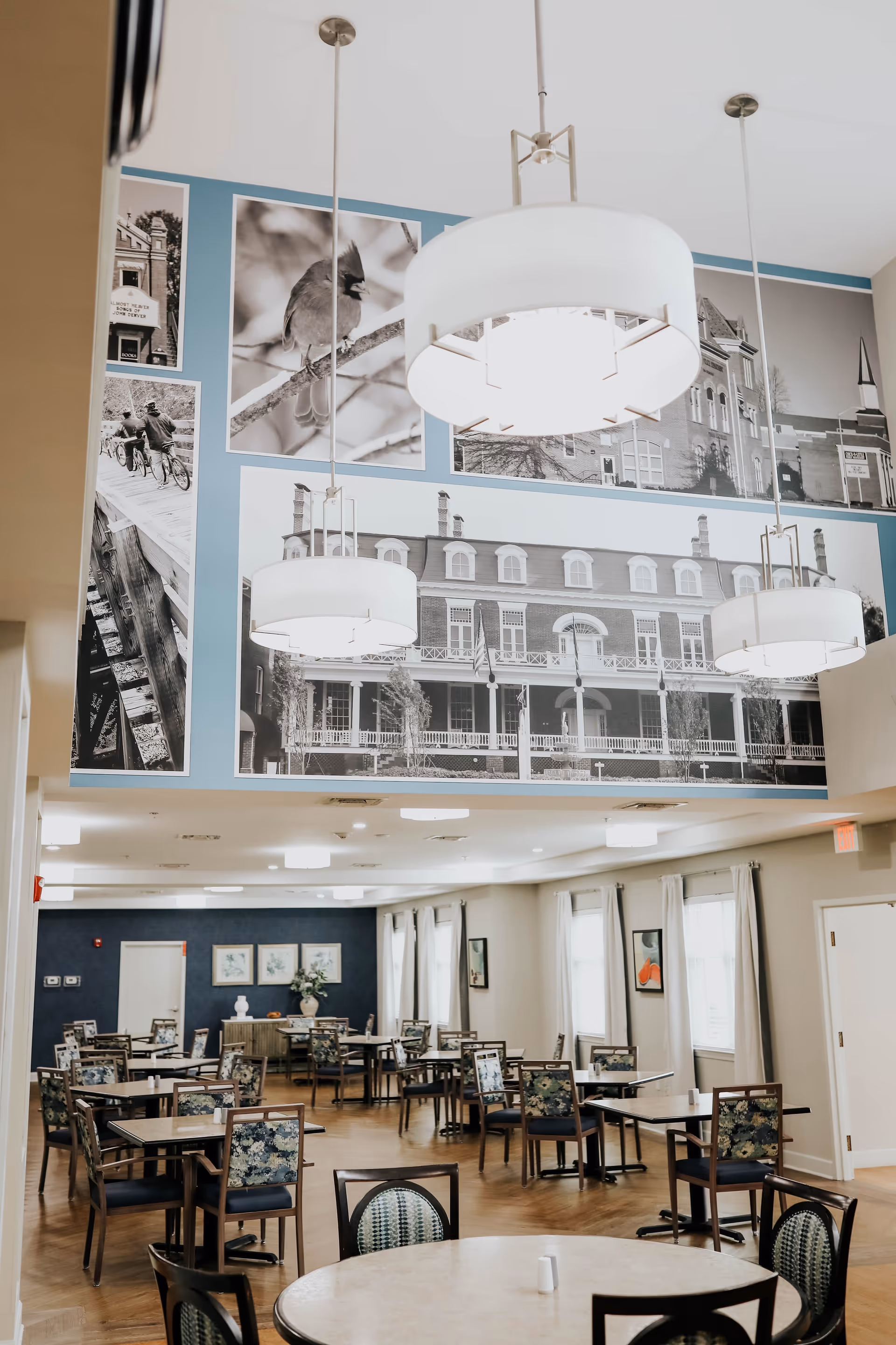 A spacious dining room with multiple tables and chairs arranged neatly. The room features large windows with curtains, modern hanging light fixtures, and a wall decorated with large black and white photographs of buildings and a bird.