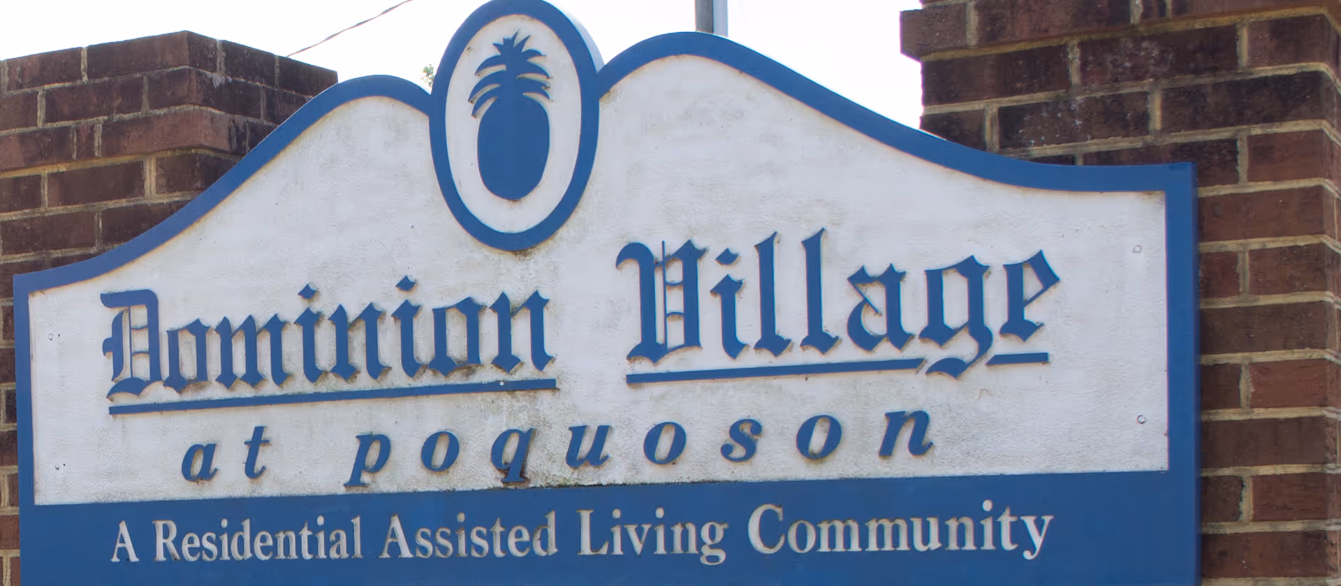 A large outdoor sign made of brick and wood displaying the name 'Dominion Village at Poquoson' with a pineapple emblem at the top. The sign also states 'A Residential Assisted Living Community.'