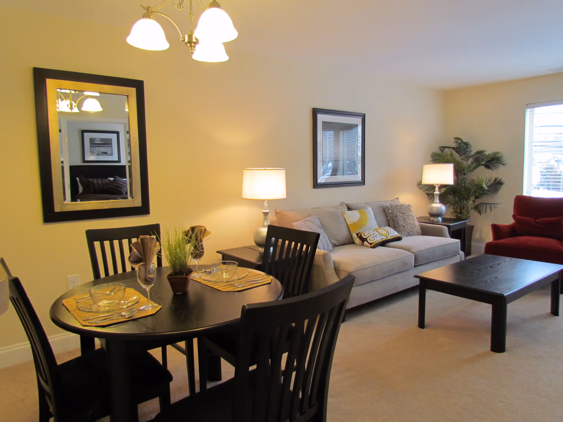 A cozy living room and dining area with a round black dining table set for two, a beige sofa with decorative pillows, a red armchair, two table lamps, a large mirror on the wall, a framed picture, and a potted plant near a window with blinds.