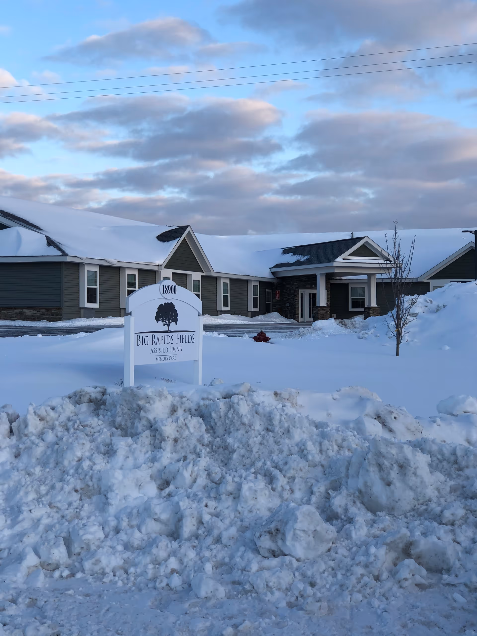 Exterior view of Big Rapids Fields Assisted Living & Memory Care facility during winter, with snow covering the ground and roof. A white sign with the facility's name and address is visible in front of the building.