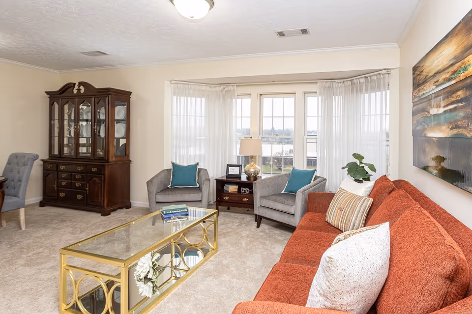 Bright living room with a red sofa, two gray armchairs, a glass-and-gold coffee table, a china cabinet, and a bay window.