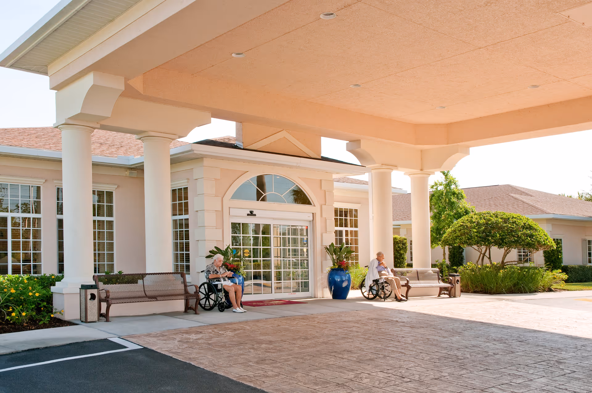 Covered front entrance of a senior living facility with columns, benches, planters, and two people in wheelchairs near the glass doors.