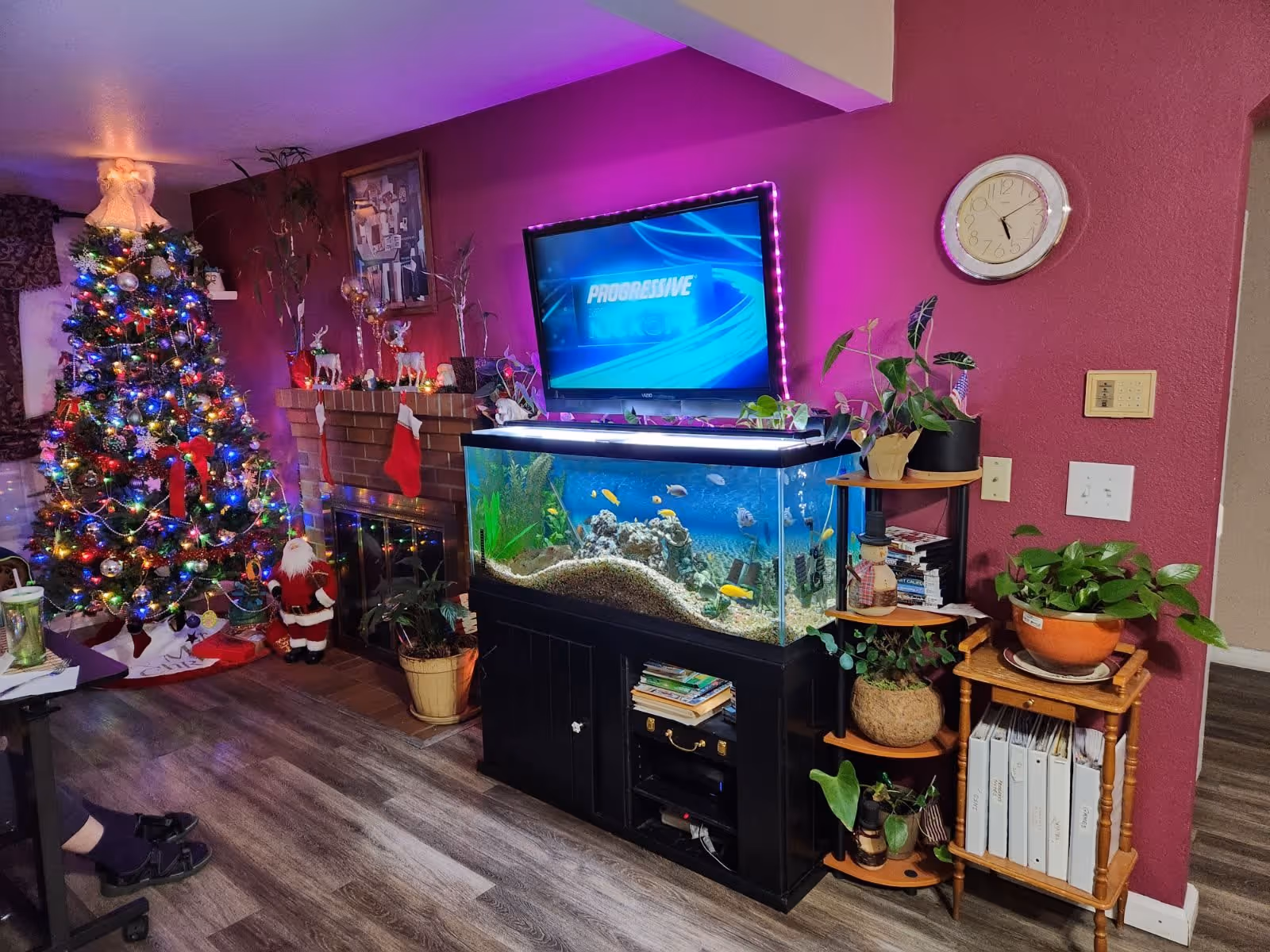 A cozy living room decorated for Christmas with a brightly lit Christmas tree, a Santa Claus figure, and stockings hanging on a brick fireplace. A large fish tank sits on a black cabinet beneath a wall-mounted TV with purple LED backlighting. Several potted plants and shelves with books and decorations are arranged around the fish tank. The walls are painted a deep red color and the floor is wood laminate.