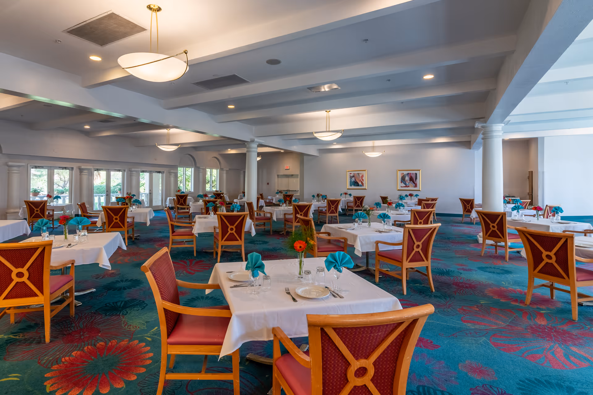 A spacious dining room with multiple tables covered in white tablecloths, each set with plates, glasses, silverware, and teal napkins folded in a decorative manner. The room features wooden chairs with red cushions and a colorful carpet with floral patterns. Large windows line one side of the room, allowing natural light to enter, and there are ceiling lights and columns throughout the space.