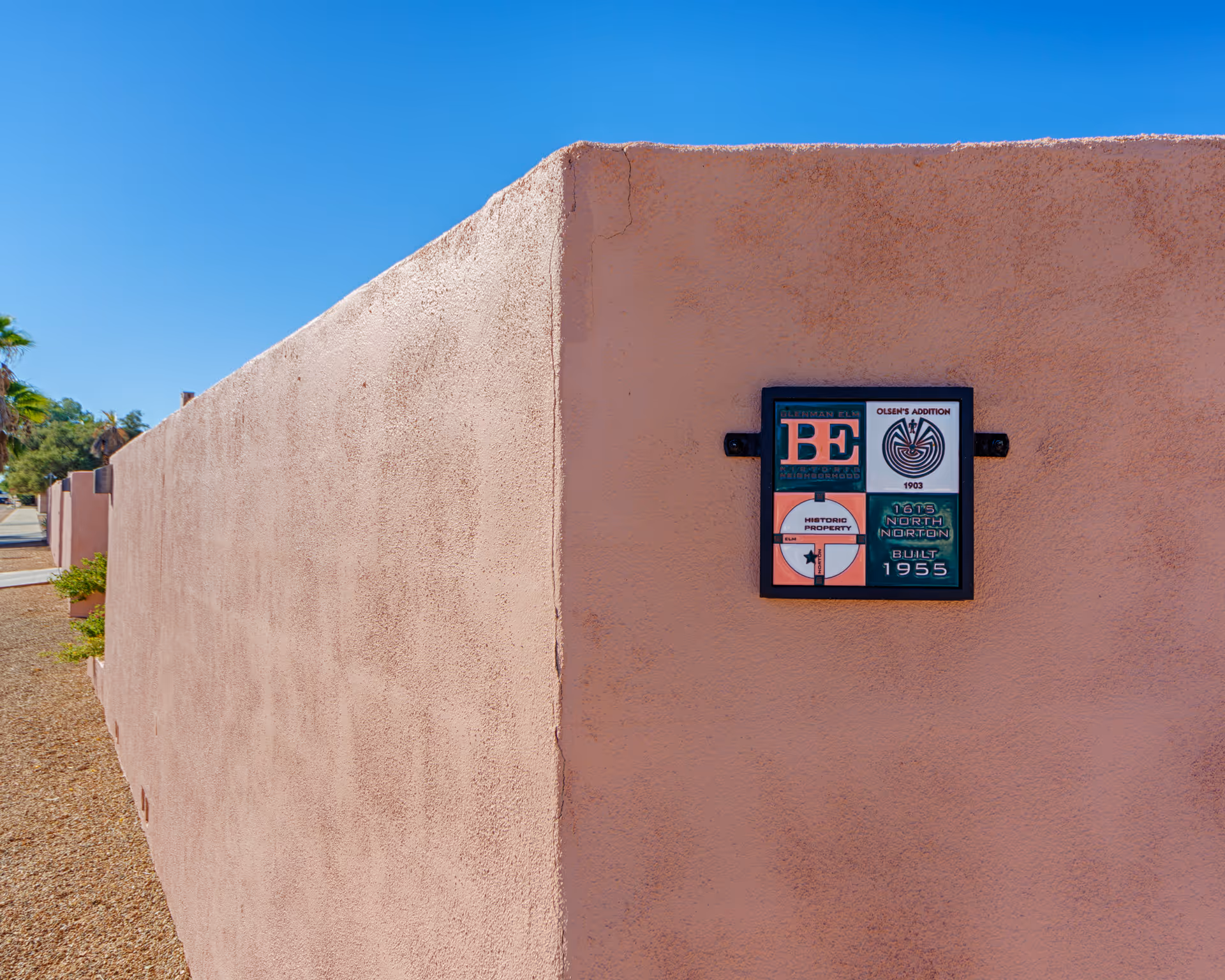 Exterior view of a peach-colored stucco wall with a small plaque mounted on it. The plaque contains multiple sections with text and symbols, including 'Blenman Elm Neighborhood', 'Olsen's Addition 1903', 'Historic Property', and '1615 North Norton Built 1955'. The sky is clear and blue, and there are some trees and plants visible along the sidewalk beside the wall.