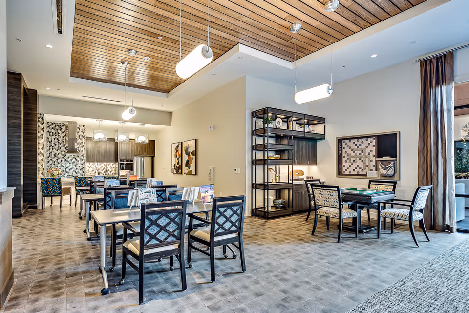 Spacious bright communal dining and activity room with several tables and chairs, pendant lighting, and an open kitchen area with patterned backsplash.