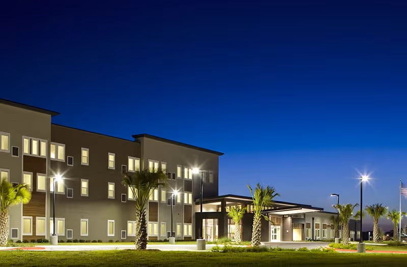 Exterior view of a modern, three-story senior living facility building at dusk with illuminated windows and street lamps, surrounded by palm trees and a well-maintained lawn.