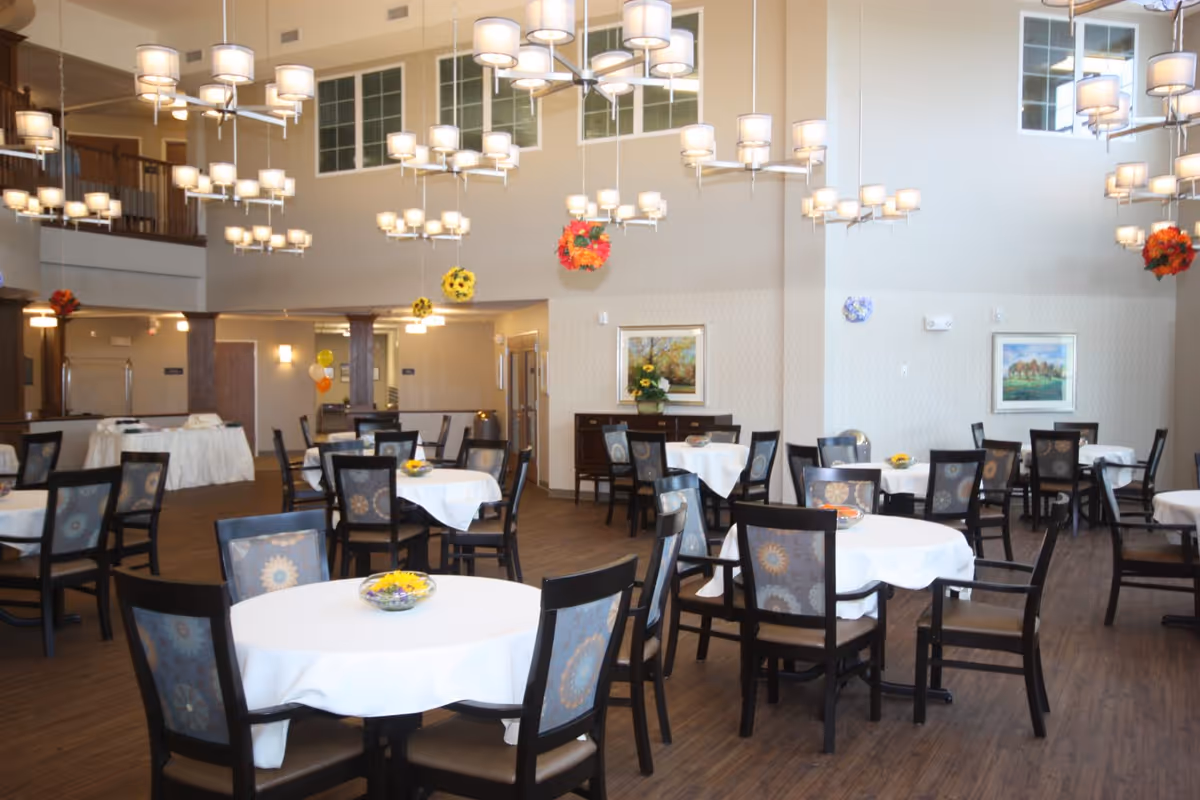 A spacious dining room with multiple round tables covered with white tablecloths, each surrounded by dark wooden chairs with patterned upholstery. The room features high ceilings with modern chandeliers hanging down, and decorative floral arrangements hanging from the ceiling. The walls are light-colored with framed artwork and a sideboard with a flower arrangement. The floor is wooden, and the overall atmosphere is bright and welcoming.