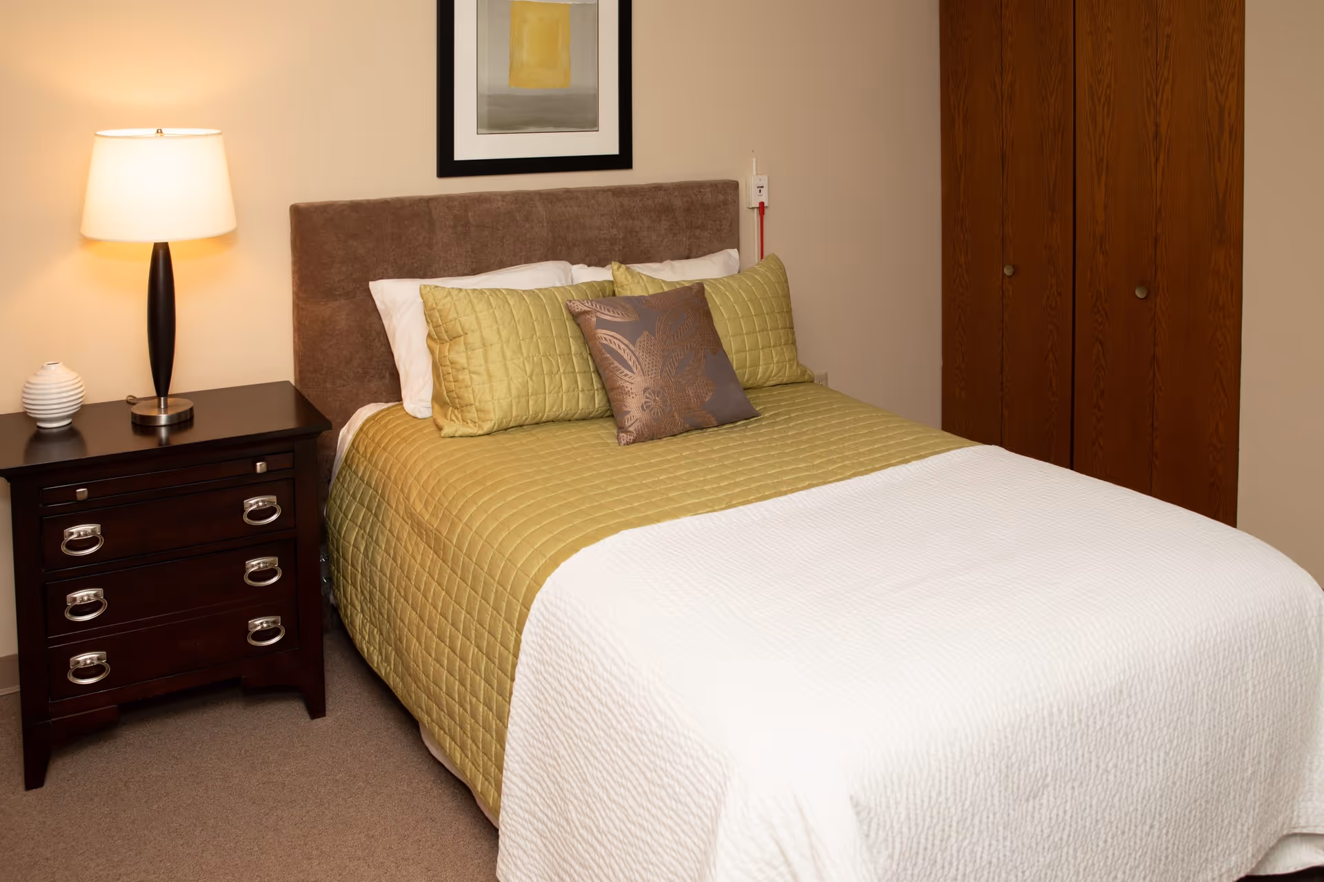 A neatly made bed with a green quilt and decorative pillows beside a dark wooden nightstand with a lamp in a small bedroom.