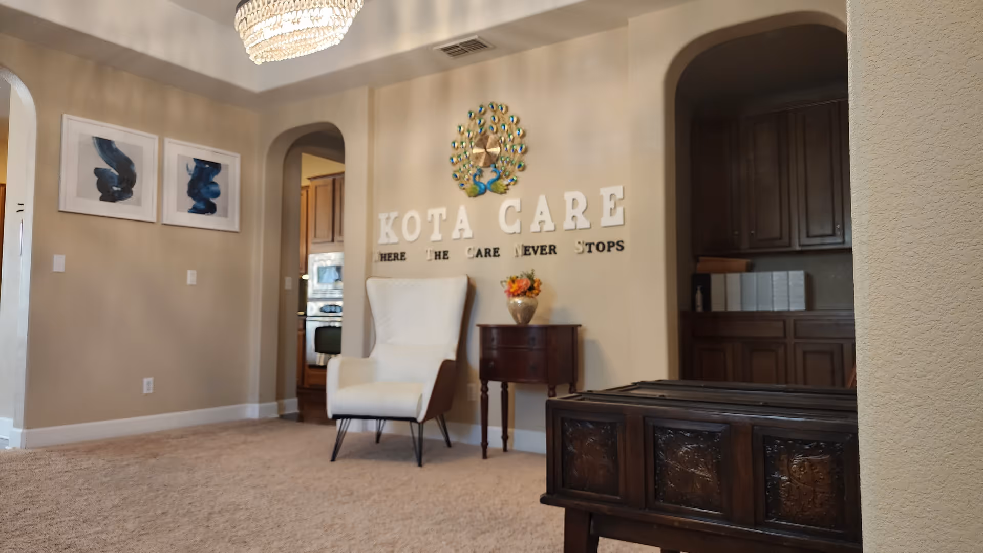 A cozy common area showing a white armchair and small table beneath wall letters reading 'KOTA CARE' with a decorative peacock and chandelier.