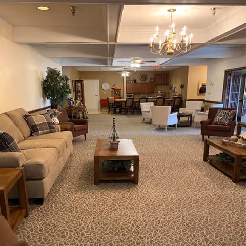 A spacious and well-lit common area in an assisted living facility featuring beige carpet with a geometric pattern, multiple seating options including sofas and armchairs, wooden coffee tables, a chandelier, and a kitchen area with dining tables and chairs in the background.