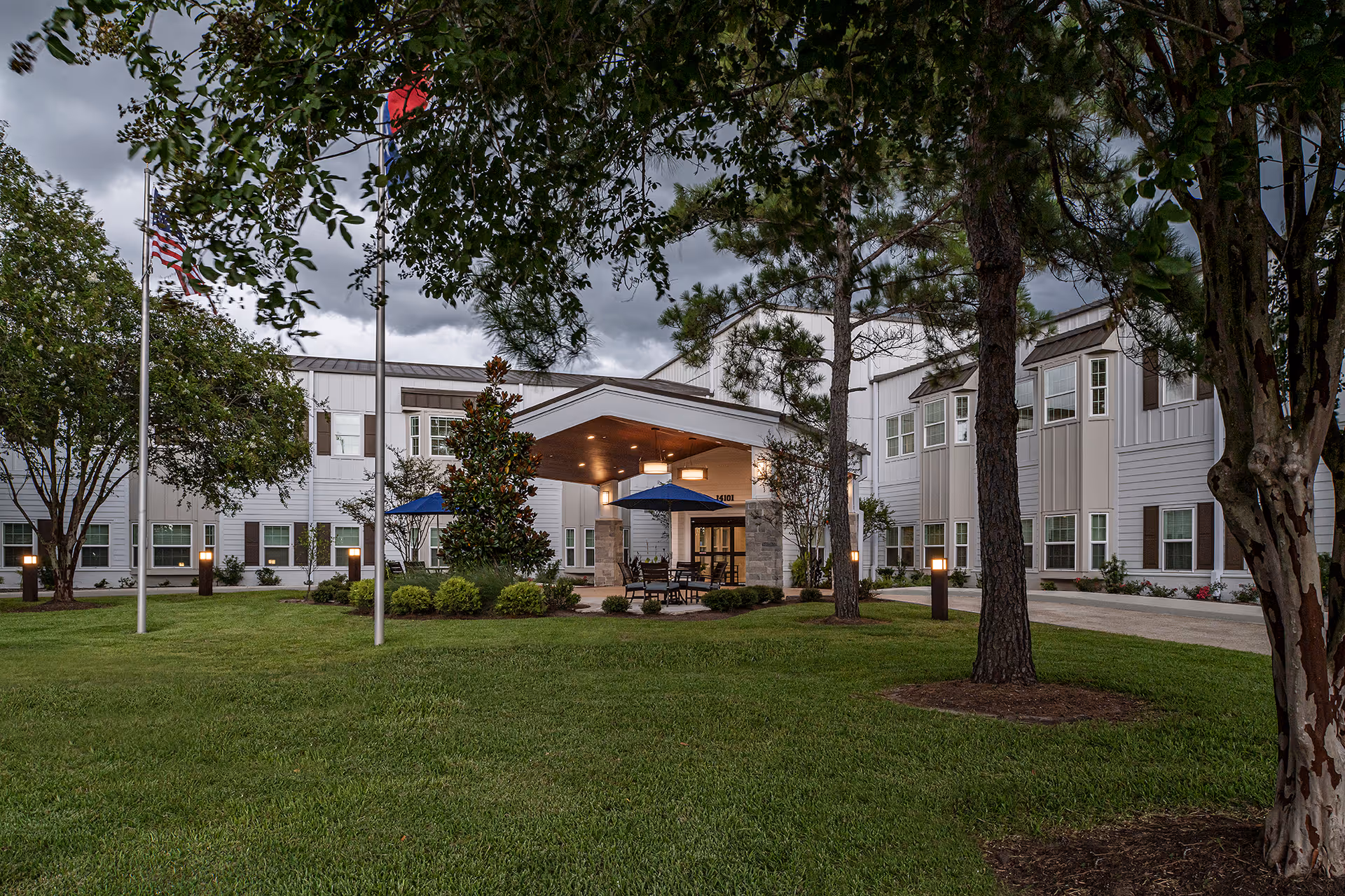 Exterior view of a senior living facility building with a covered entrance, surrounded by green grass, trees, and flagpoles. The sky is cloudy and the building has white siding with multiple windows.