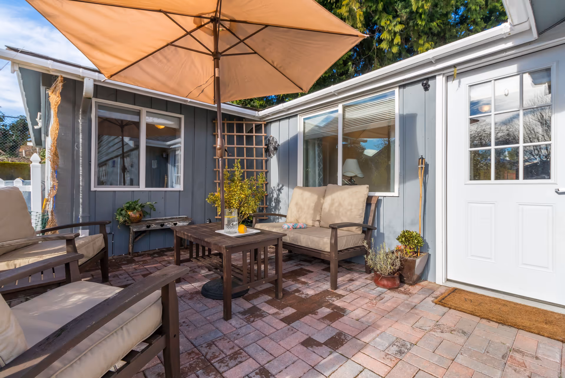 Outdoor patio with beige cushioned seating, a large umbrella, wooden coffee table, potted plants and a white door beside a gray house on a brick-paved area.