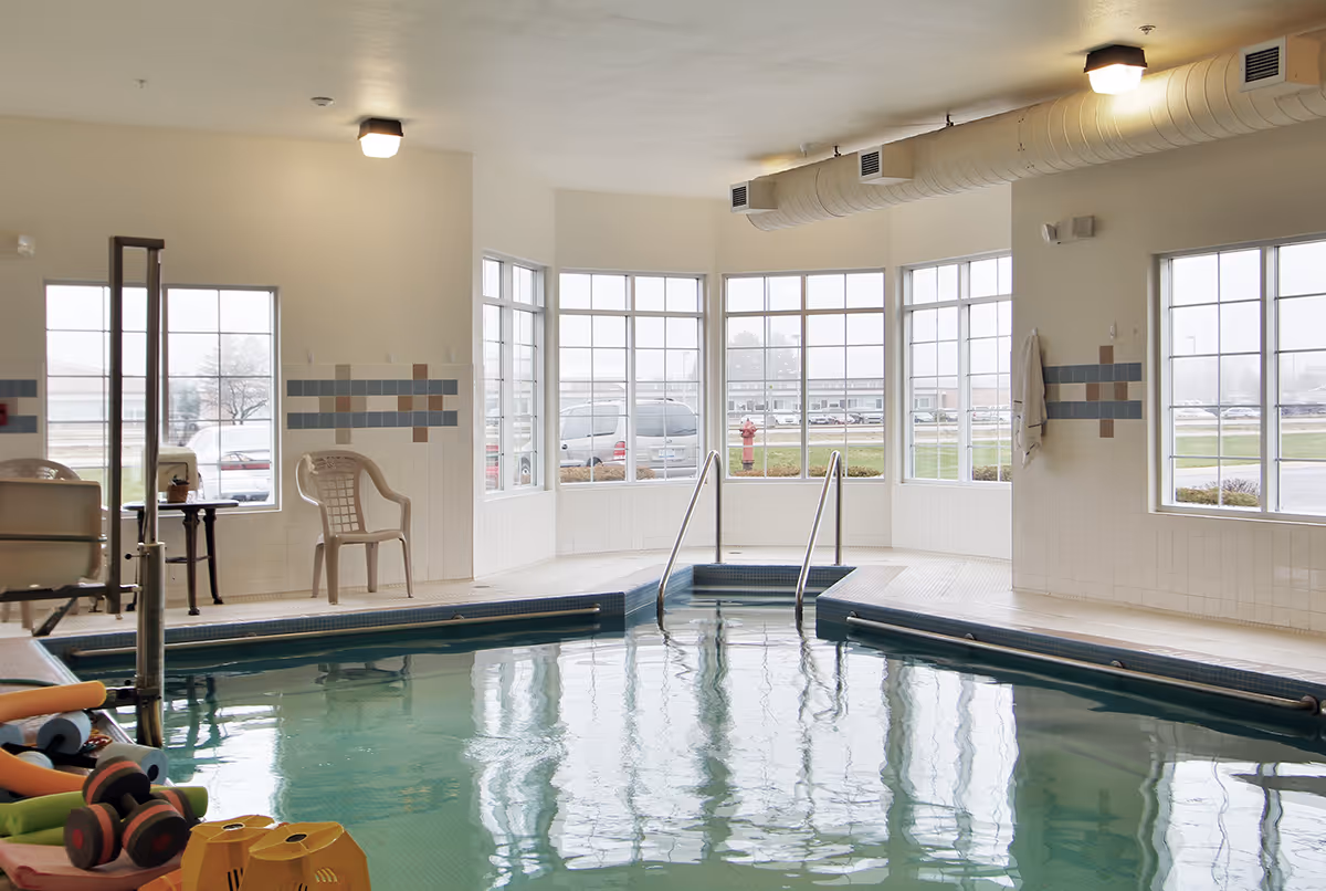 Indoor swimming pool area with large windows allowing natural light to enter. The pool has handrails and steps leading into the water. There are plastic chairs and pool equipment such as dumbbells and foam noodles around the pool. The walls are tiled with a decorative pattern and the ceiling has exposed ventilation ducts.