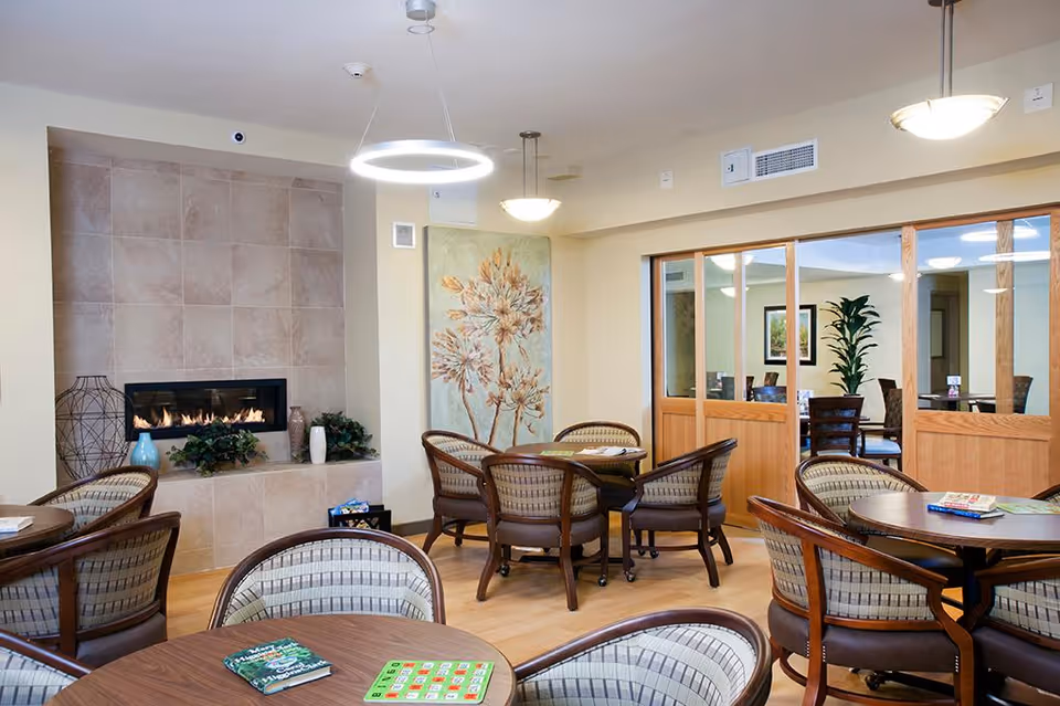 A cozy common area with round tables and cushioned chairs arranged around a modern fireplace set in a tiled wall. The room features soft lighting from ceiling fixtures, a large floral painting on the wall, and a wooden sliding door leading to another room with additional seating and plants.