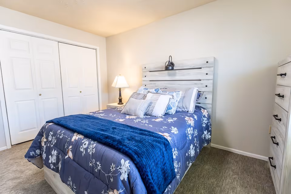A bedroom with a bed featuring a floral blue comforter and multiple pillows. The bed has a light wooden headboard with a mounted reading light. There is a nightstand with a lamp on the left side of the bed, a white double-door closet in the background, and a matching light wooden dresser on the right side. The walls are painted white and the floor is carpeted.