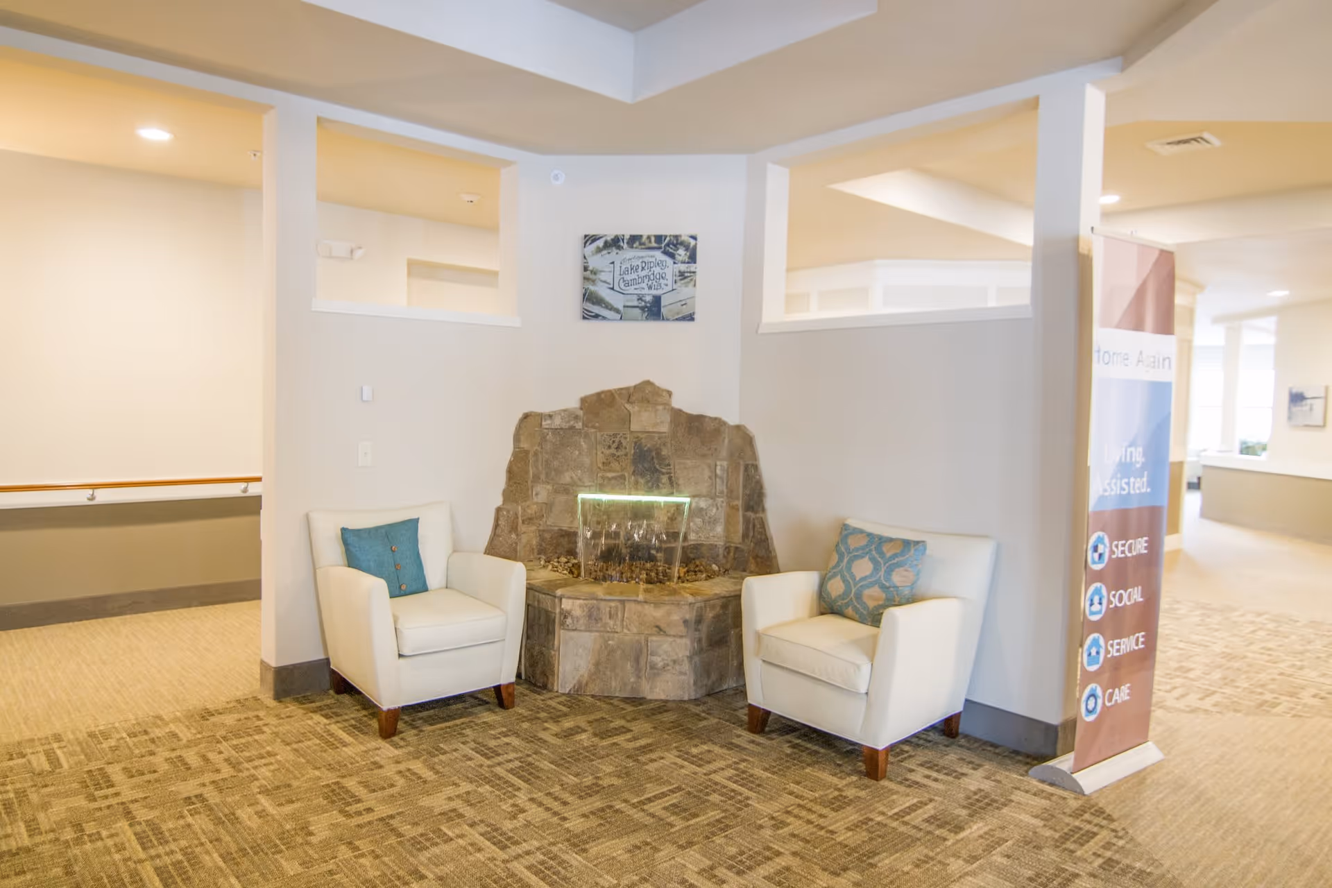 A cozy seating area in a senior living facility featuring two white armchairs with decorative pillows placed on either side of a small stone water fountain. The area has beige walls, carpeted flooring, and a banner on the right side listing features like Secure, Social, Service, and Care.