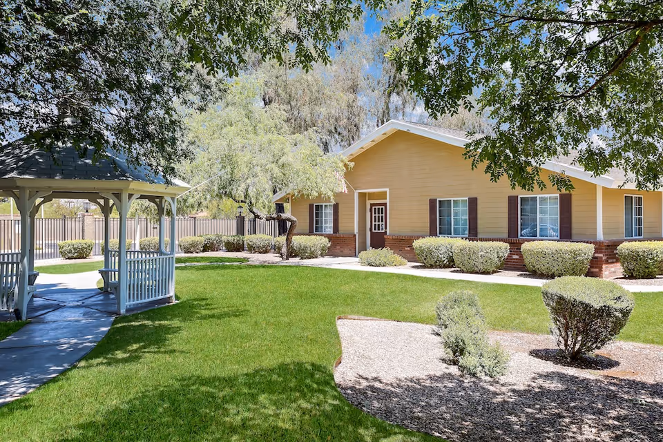 A sunny outdoor garden area at Tucson Gardens Memory Care featuring a well-maintained green lawn, trimmed bushes, a white gazebo with a paved walkway, and a yellow building with brown shutters in the background. Trees provide shade over part of the scene.