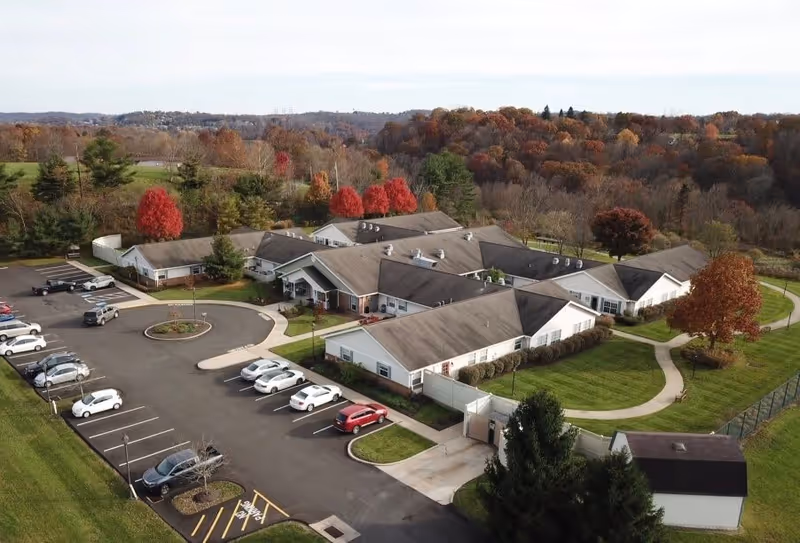 Aerial view of Arden Courts - ProMedica Memory Care Community in Jefferson Hills, showing multiple single-story buildings with gray roofs surrounded by green lawns and trees with autumn foliage. There is a parking lot with several cars parked and a curved driveway leading to the main entrance. The background features a wooded area with colorful fall trees.