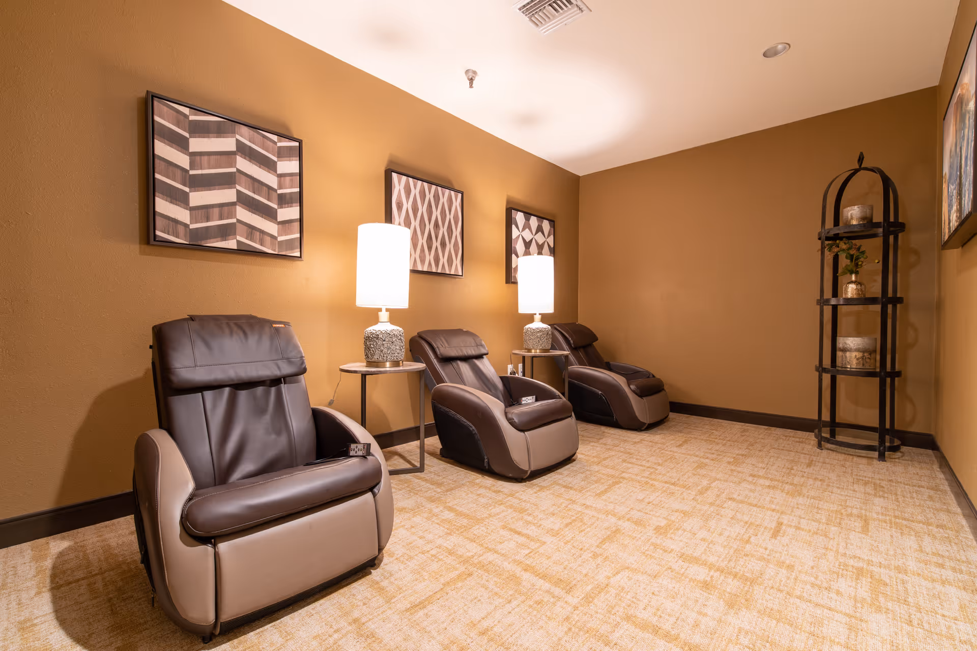 Three leather recliner massage chairs in a warmly lit relaxation room with side tables, lamps, framed wall art, and a decorative shelf.