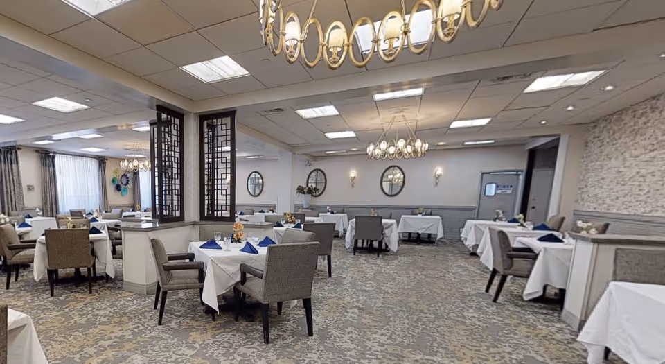 A spacious dining room with multiple tables covered in white tablecloths, each set with blue napkins and small floral centerpieces. The room features beige patterned carpeting, cushioned chairs, decorative wall mirrors, and elegant chandeliers hanging from the ceiling. Light filters in through large windows with curtains on the left side.