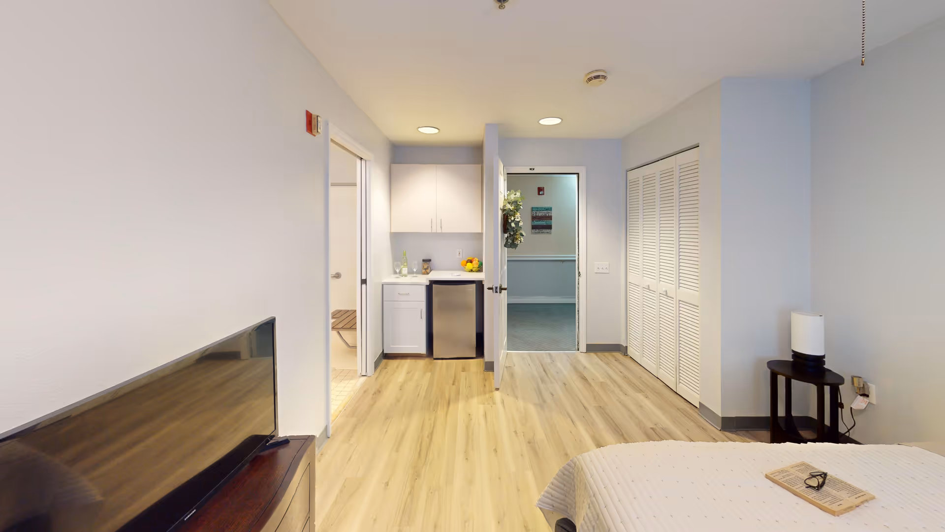 Interior view of a senior living facility room with light wood flooring, a bed partially visible on the right, a flat-screen TV on a wooden stand on the left, a small kitchenette with white cabinets and a mini fridge in the center background, and an open door leading to a hallway. There is a small table with a lamp next to the bed and a closet with white louvered doors on the right wall.