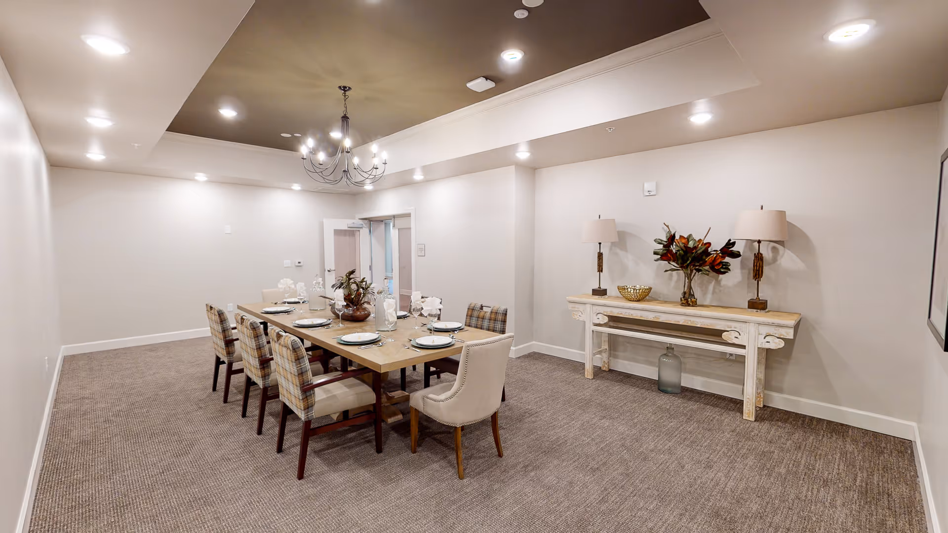 A dining room with a long wooden table set for eight people with plates, glasses, and napkins. The chairs around the table have a mix of plaid and solid upholstery. The room has beige walls, carpeted floor, recessed lighting, and a chandelier hanging from the ceiling. Against one wall is a console table with two lamps, a decorative bowl, and a vase with flowers.