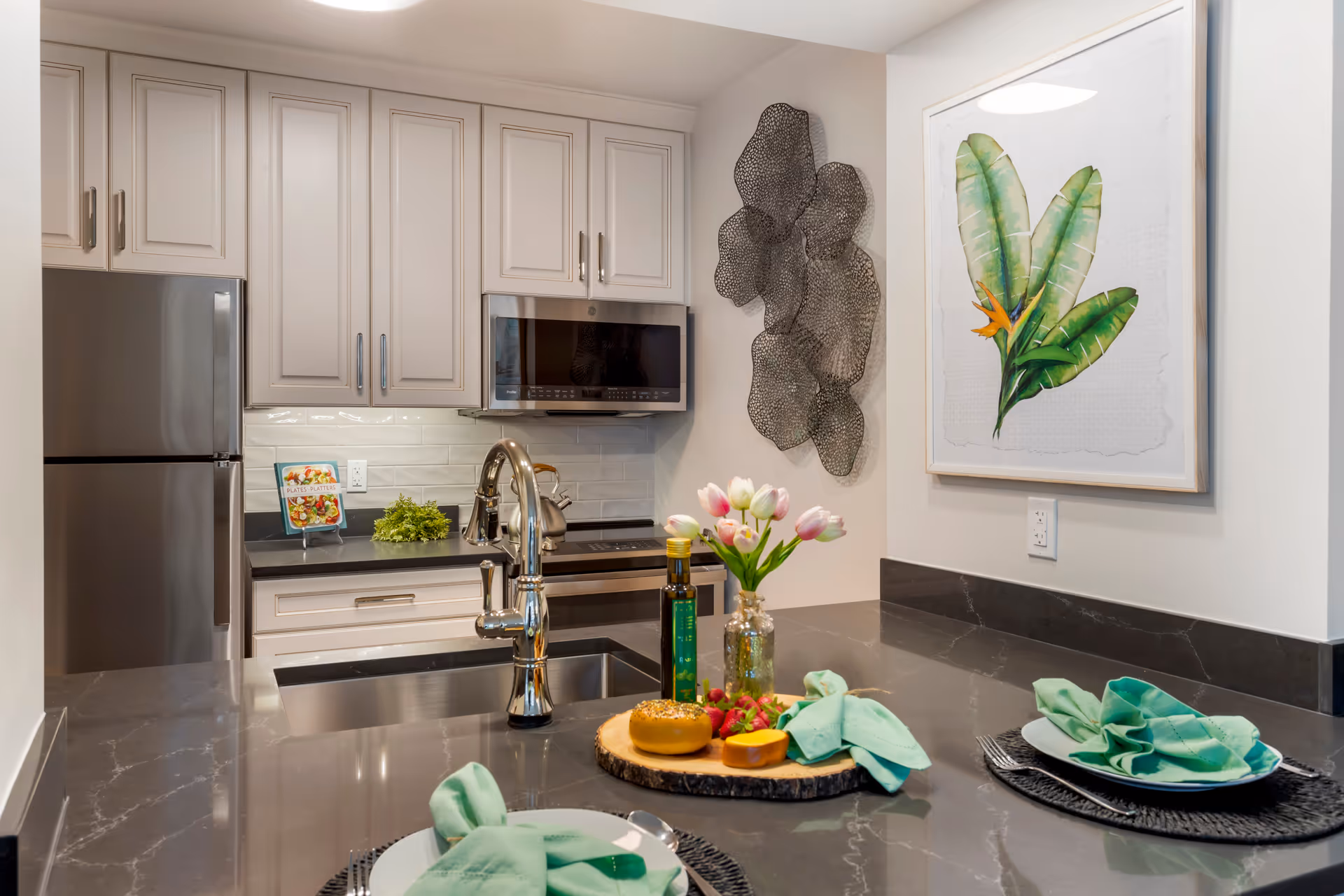 Modern kitchen with gray countertops and white cabinets. Stainless steel refrigerator and microwave are visible. A kitchen island with a sink has two place settings with green napkins, plates, and cutlery. A wooden tray with a bagel, cheese, strawberries, two bottles, and a vase with pink tulips is on the island. Wall decor includes a metal art piece and a framed botanical print.