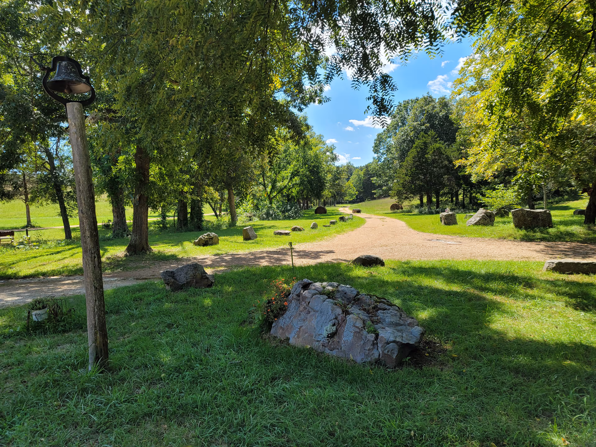 A sunlit grassy outdoor area with a winding dirt path, scattered large rocks and trees, and a wooden lamp post in the foreground.