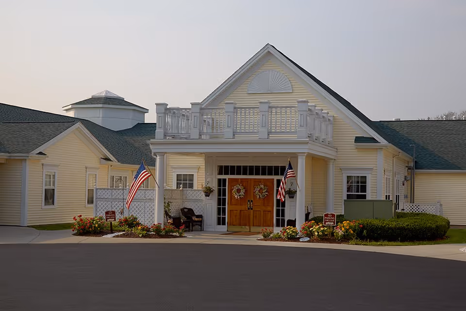 Front entrance of a pale yellow senior living building with a covered porch, double wooden doors, American flags, and landscaping.