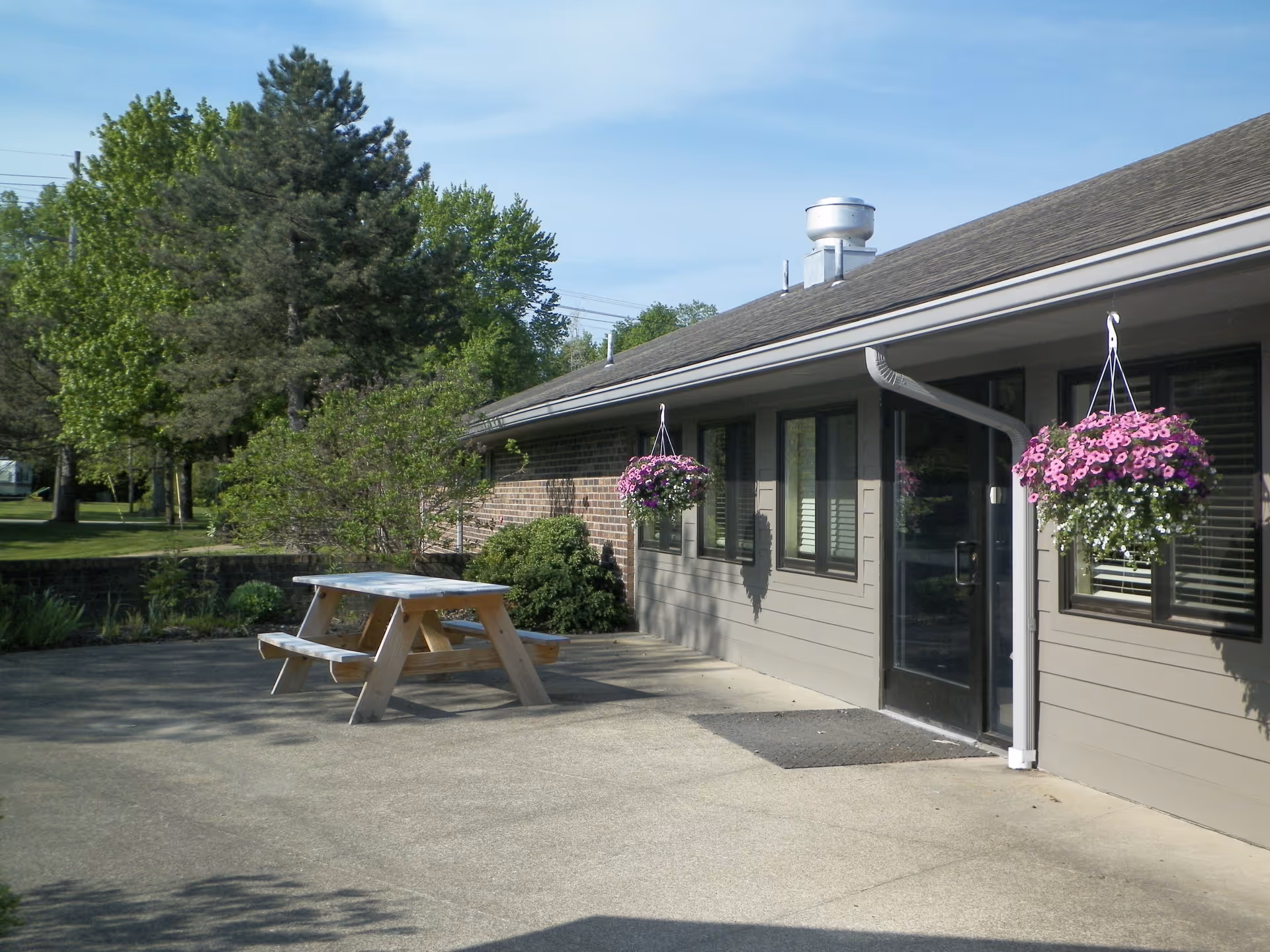 Exterior patio of a single-story building with a picnic table, hanging flower baskets, and trees by a small pond.