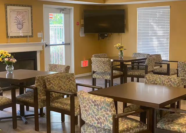 Interior view of a senior living facility common area with multiple square tables and cushioned chairs featuring a leaf pattern. There is a fireplace with a framed artwork above it on the left wall, a glass door leading outside, a wall-mounted TV, and windows with blinds. Small vases with yellow flowers are placed on some tables.