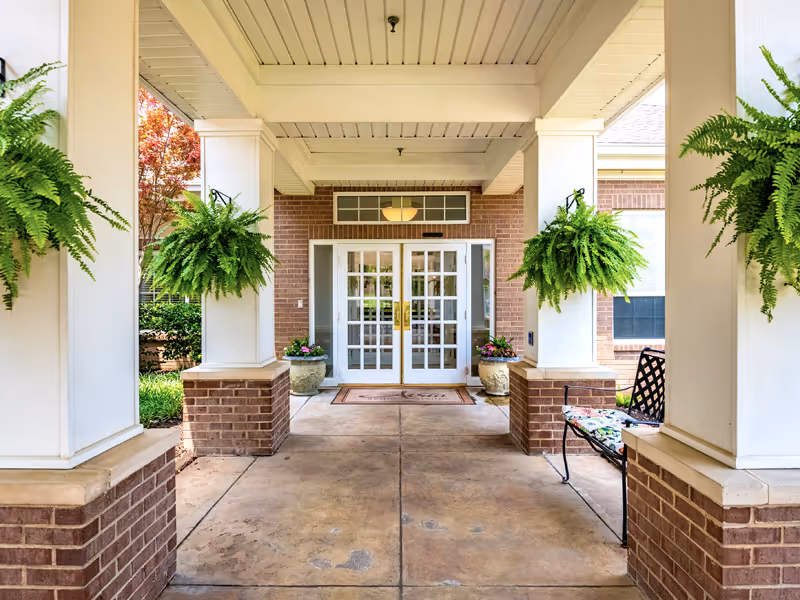 This image shows a covered brick entrance with white pillars, hanging ferns, and double glass doors.