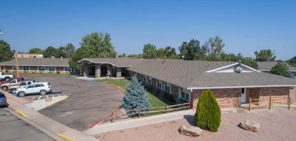 Front exterior of a single-story brick senior care building with a covered entrance, parking lot, and landscaped gravel and shrub areas.