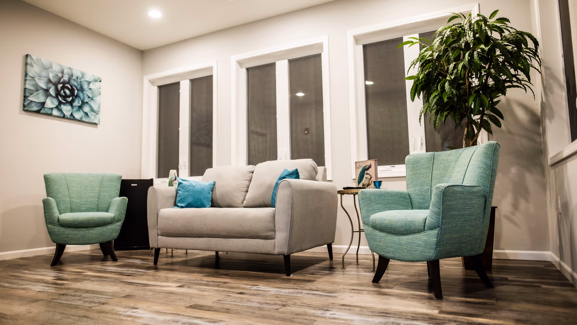 A cozy living room area with a light gray loveseat adorned with blue throw pillows, flanked by two teal armchairs. The room features three large windows with closed blinds, a small side table with decorative items, a large potted plant, and a wall painting of a succulent. The floor is wooden with a natural finish.